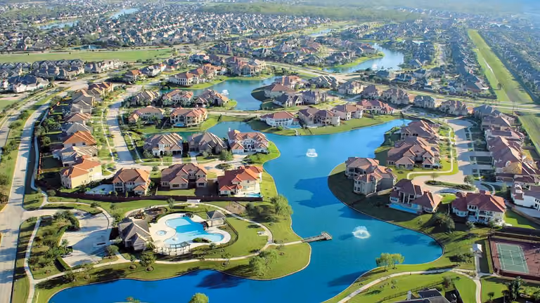 Aerial photograph of houses around a chain of lakes with fountains: large houses with red and brown roofs line the curving shores, and a small pier reaches into the water from a lakeside path. A pool and pool house sit at lower left and a tennis court at lower right. Beyond them, more lakes and streets of houses run to the horizon.